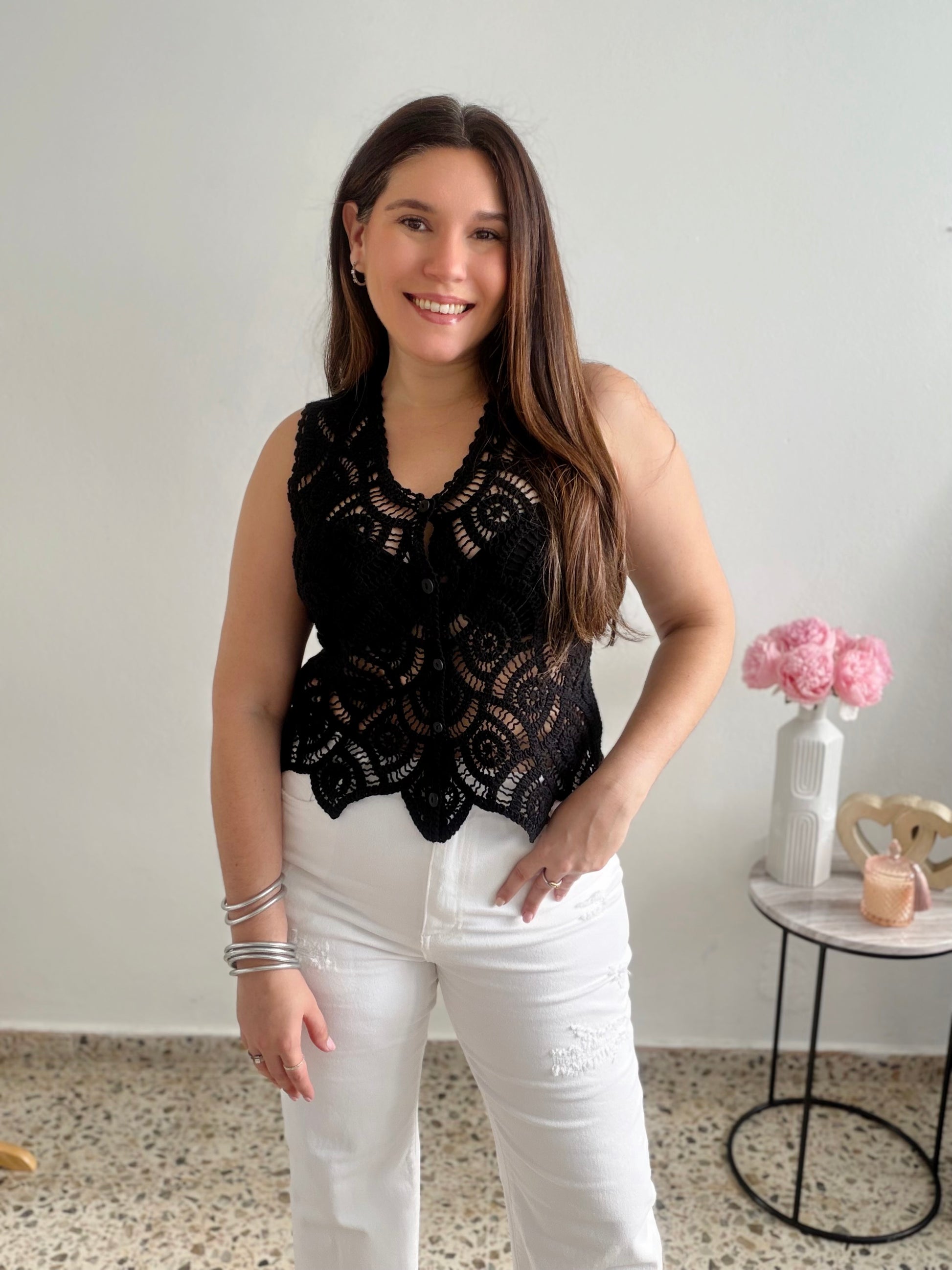 Woman wearing a black lace top and white pants standing in a room with a small table and flowers.