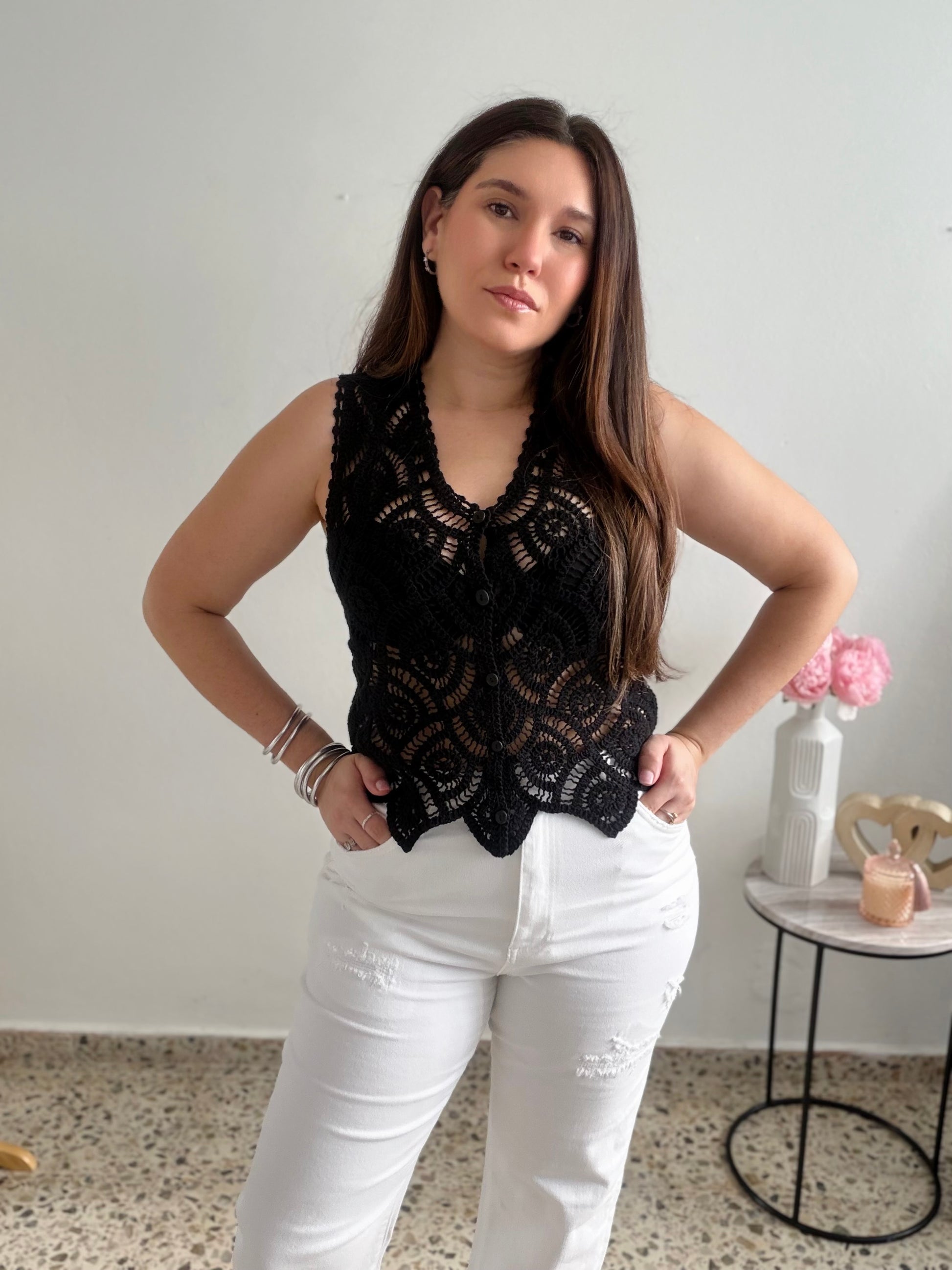 Woman wearing a black crochet top and white pants standing in a room with a small table and vase in the background.